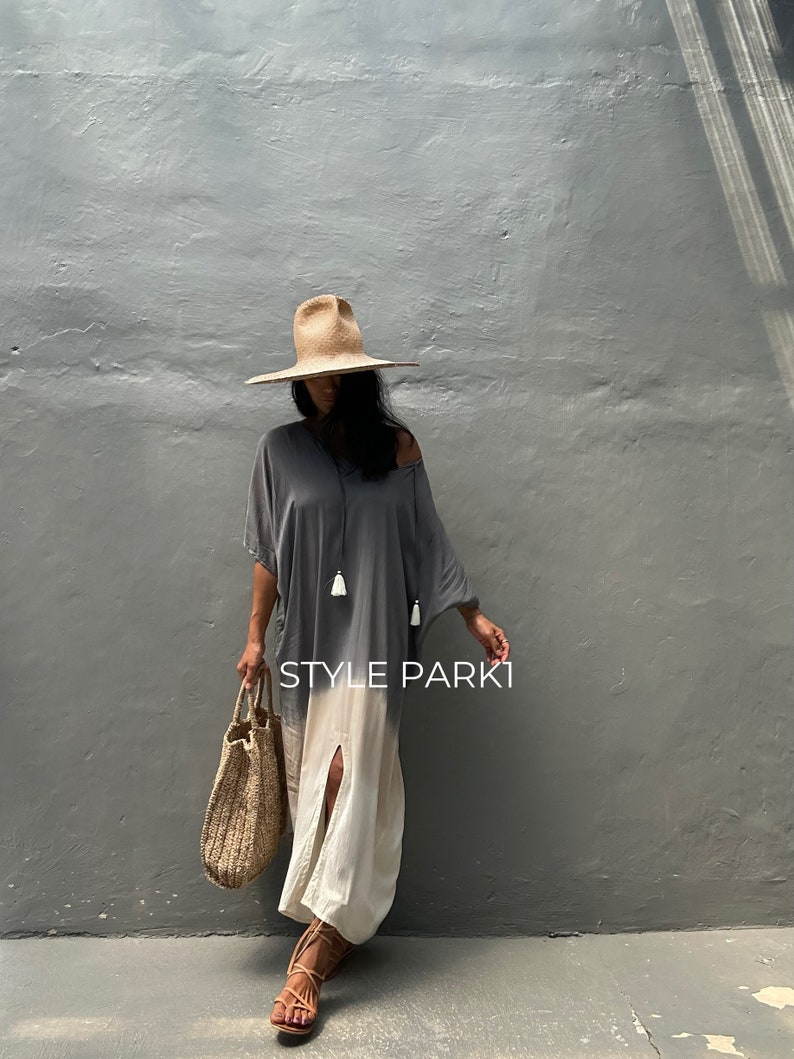May include: A woman wearing a gray and white ombre maxi dress with a slit up the front. She is also wearing a wide-brimmed straw hat, a woven tote bag, and brown sandals. The text "STYLE PARKI" is visible on the dress.