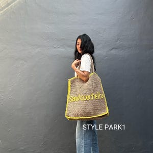 May include: A natural-fibre tote bag with the text "sand coachella" in yellow. The bag has a yellow trim. The bag is carried by a person wearing a white shirt and blue jeans. The bag is made of a natural fiber.