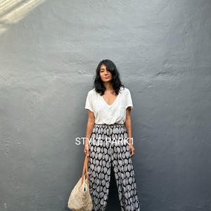May include: A woman wearing a white V-neck shirt and black and white patterned pants. She is holding a straw tote bag with a leather handle and wearing sandals. The text "STYLE PARK1" is visible on the pants.