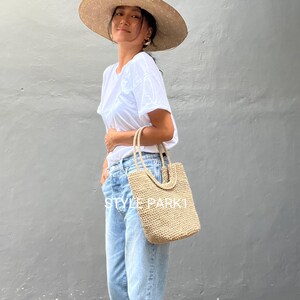 Pu&ograve; includere: Una donna indossa una maglietta bianca, jeans blu chiaro e un cappello di paglia a tesa larga e tiene una borsa tote in tessuto beige. La borsa ha un design unico con un motivo intrecciato.