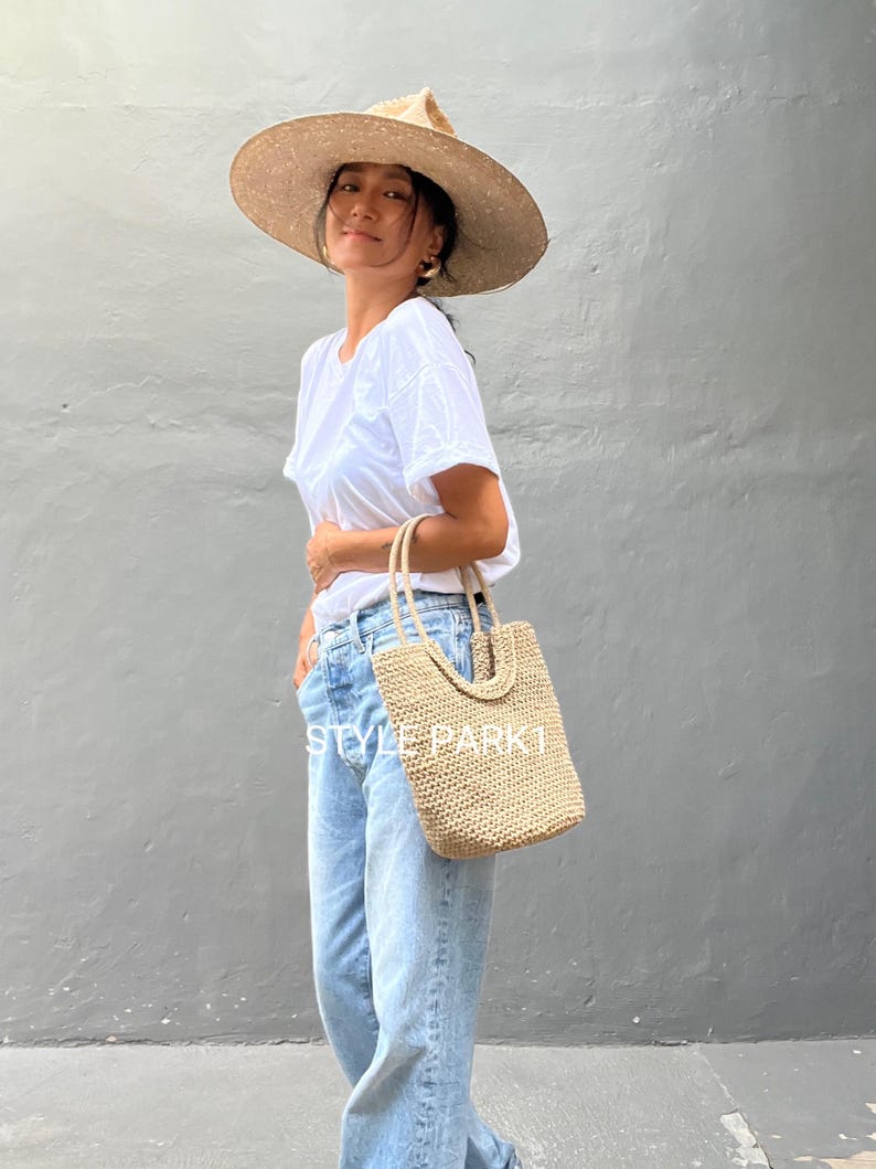 Pu&ograve; includere: Una donna indossa una maglietta bianca, jeans blu chiaro e un cappello di paglia a tesa larga e tiene una borsa tote in tessuto beige. La borsa ha un design unico con un motivo intrecciato.