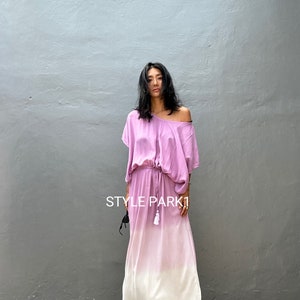 Puede incluir: Una mujer lleva un vestido largo degradado de rosa a blanco. El vestido tiene un corte holgado y una cintura con cordón. El vestido está hecho de una tela suave y fluida. La mujer está de pie frente a una pared gris. El texto "STYLE PARK1" es visible en la pared.