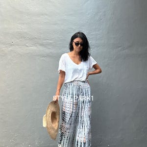 May include: A woman wearing a white t-shirt and tie-dye wide-leg pants. She is holding a straw hat and wearing sunglasses. The text "STYLE PARK1" is visible in the image.