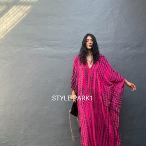 May include: A floor-length kaftan dress in vibrant fuchsia pink featuring a vertical dark charcoal tie-dye pattern, a deep V-neckline, and wide flowing sleeves. The text STYLE PARK1 is printed in white on the front of the garment.