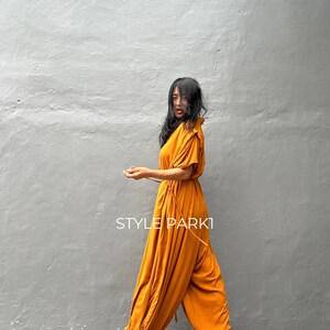 May include: A woman wearing a mustard yellow jumpsuit with a cinched waist and a ruffled neckline. The jumpsuit has a wide leg and is made of a soft, flowing fabric. The woman is standing in front of a gray wall. The text "STYLE PARK1" is visible on the image.