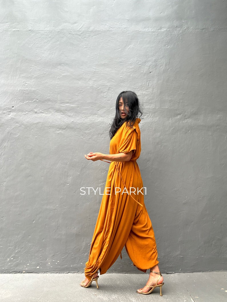 May include: A woman wearing a mustard yellow jumpsuit with a cinched waist and a ruffled neckline. The jumpsuit has a wide leg and is made of a soft, flowing fabric. The woman is standing in front of a gray wall. The text "STYLE PARK1" is visible on the image.