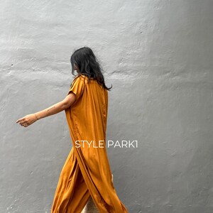May include: A woman wearing a mustard yellow jumpsuit with a loose, flowing fit. The jumpsuit has a gathered detail at the waist and a wide leg. She is carrying a woven straw tote bag and wearing gold sandals.