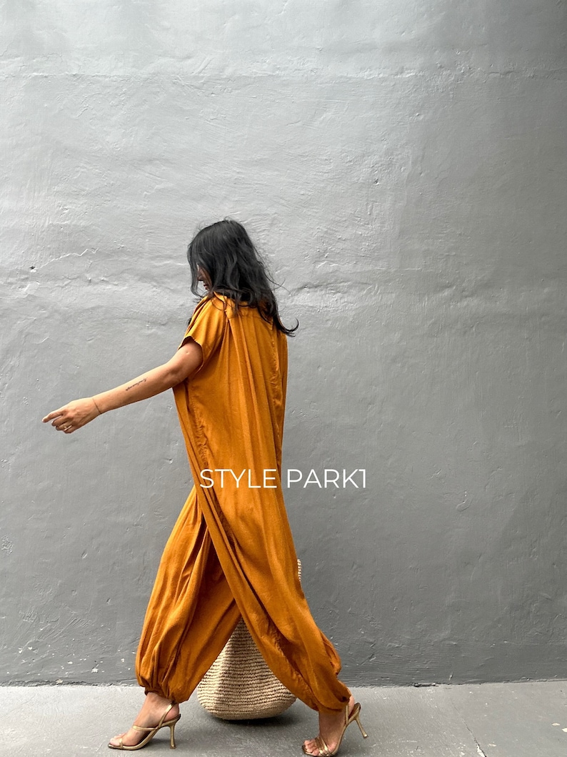 May include: A woman wearing a mustard yellow jumpsuit with a loose, flowing fit. The jumpsuit has a gathered detail at the waist and a wide leg. She is carrying a woven straw tote bag and wearing gold sandals.