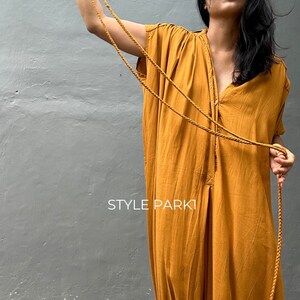 May include: A mustard yellow jumpsuit with a button-up front and a long, flowing silhouette. The jumpsuit has a relaxed fit and is made of a lightweight fabric. The model is wearing a long, brown cord necklace.