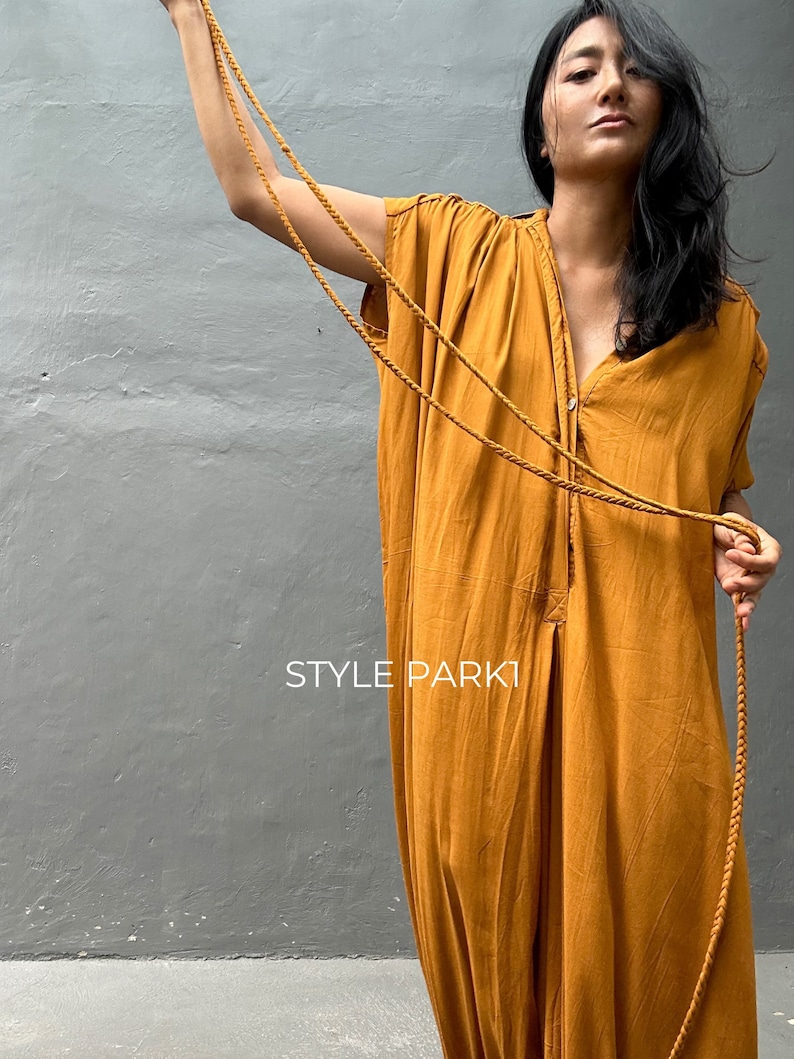 May include: A mustard yellow jumpsuit with a button-up front and a long, flowing silhouette. The jumpsuit has a relaxed fit and is made of a lightweight fabric. The model is wearing a long, brown cord necklace.