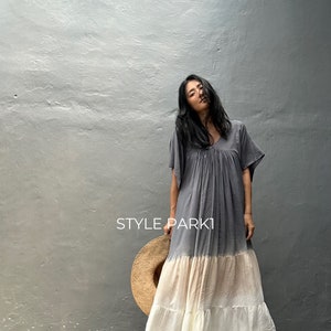 Puede incluir: Un vestido largo gris, beige y blanco con cuello en V y mangas cortas. El vestido tiene una falda escalonada y se usa con sandalias negras. Se sostiene un sombrero de paja cerca del vestido. El texto "STYLE PARK1" es visible.