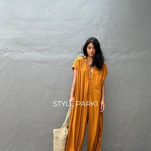 May include: A woman wearing a mustard yellow jumpsuit with a button-up front and short sleeves. She is carrying a large, woven tote bag and is standing in front of a gray wall. The text "STYLE PARK1" is visible in the image.