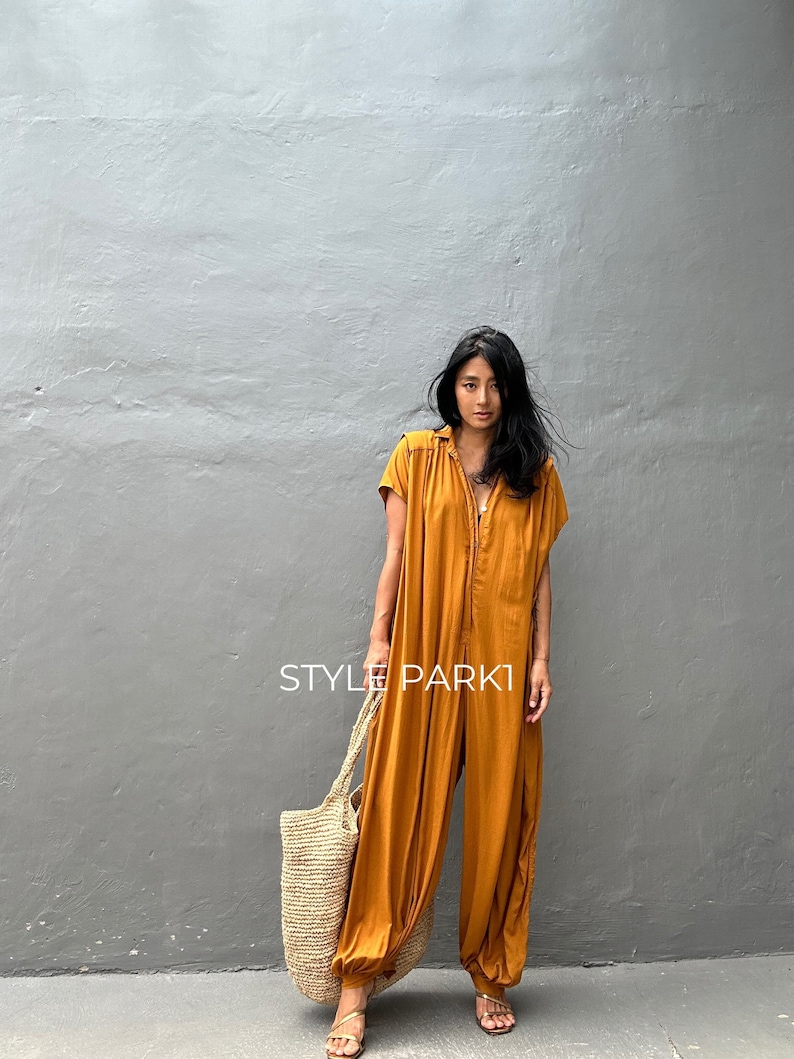 May include: A woman wearing a mustard yellow jumpsuit with a button-up front and short sleeves. She is carrying a large, woven tote bag and is standing in front of a gray wall. The text "STYLE PARK1" is visible in the image.