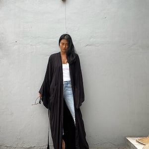 May include: A long, black kimono-style robe with wide sleeves and a tie belt. The robe is worn over a white top and blue jeans. The person is holding sunglasses.