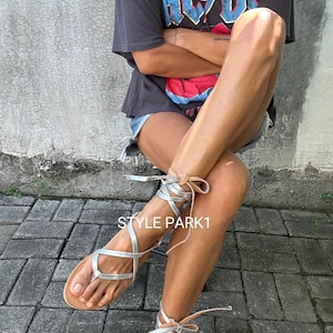 May include: Silver leather sandals with laces that wrap around the ankles. The sandals are worn by a person wearing a black t-shirt with the text "AC/DC" printed on it.