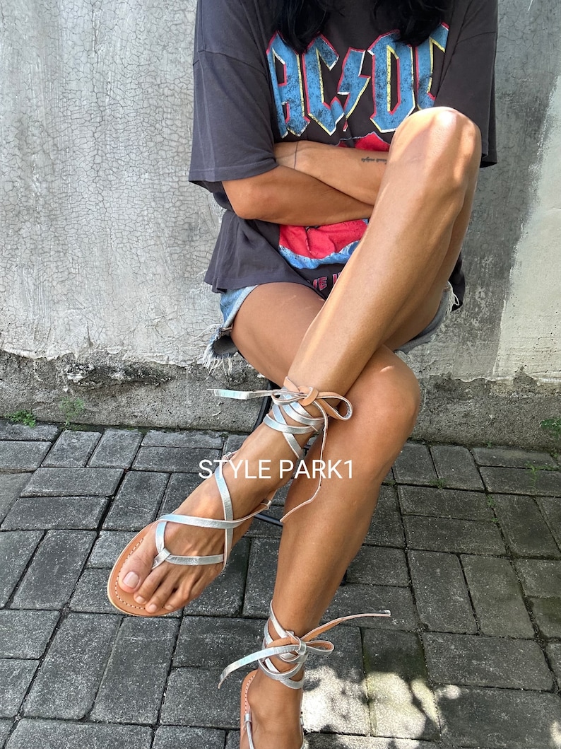 May include: Silver leather sandals with laces that wrap around the ankles. The sandals are being worn by a person wearing a black t-shirt with the text "AC/DC" printed on it.