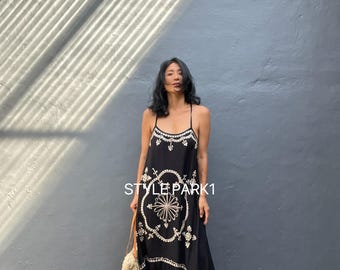 May include: A black maxi dress with white embroidered floral patterns and spaghetti straps. The dress has a tiered skirt and is worn with sandals and a woven tote bag. The text "STYLEPARK1" is visible on the dress.