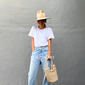 Pu&ograve; includere: Una donna indossa una maglietta bianca, jeans blu chiaro e un cappello di paglia a tesa larga. Porta una borsa a mano di paglia con la scritta "STYLE PARK1" su di essa.