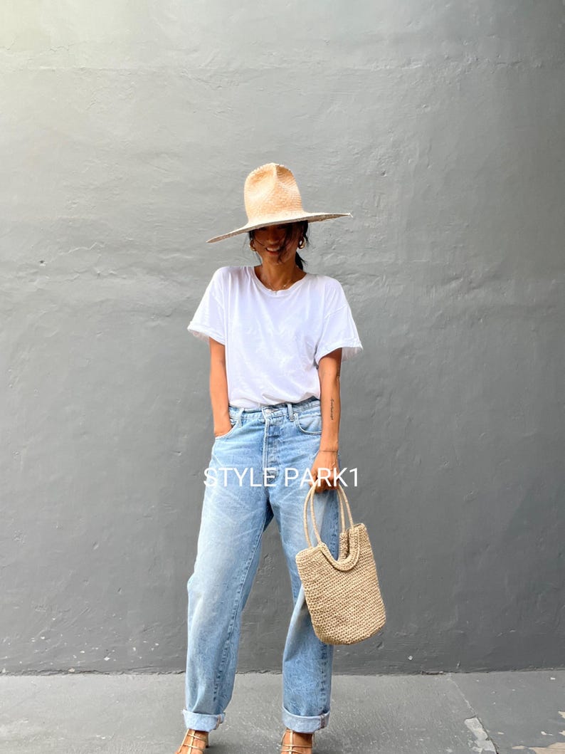 Pu&ograve; includere: Una donna indossa una maglietta bianca, jeans blu chiaro e un cappello di paglia a tesa larga. Porta una borsa a mano di paglia con la scritta "STYLE PARK1" su di essa.