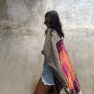 May include: A patterned kimono in black, gold, orange, and purple. The kimono features a mandala design on the back and is worn over a white top and denim shorts. The person is wearing sunglasses and carrying a woven bag.