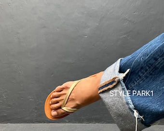 May include: A gold-colored thong sandal with a brown sole. The sandal is worn with blue denim jeans, the cuff of which is turned up. The text "STYLE PARK1" is visible on the jeans.