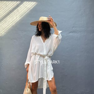 Puede incluir: Un vestido blanco de camisa de manga larga con cinturón de lazo. El vestido se lleva con un sombrero de paja y un bolso de mano tejido. El texto "STYLE PARK1" es visible en el vestido.