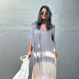 May include: A woman wearing a gray and white tie-dye maxi dress with a V-neck and tassels. She is holding a white tote bag with the word "ARK1" printed on it.