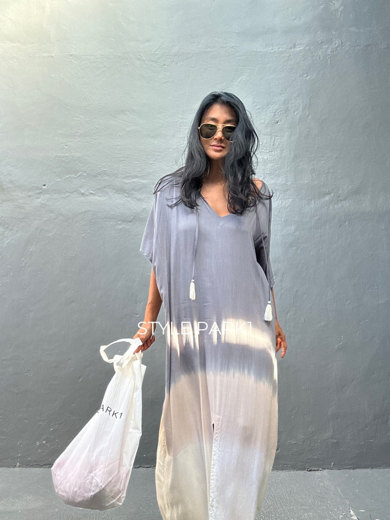 May include: A woman wearing a gray and white tie-dye maxi dress with a V-neck and tassels. She is holding a white tote bag with the word "ARK1" printed on it.