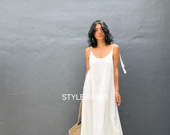 Könnte beinhalten: Ein weißes Maxikleid mit dünnen Trägern und einem Rüschen-Saum. Das Kleid hat ein schlichtes, elegantes Design und ist perfekt für einen Sommertag. Das Model trägt das Kleid mit einer Strohtasche und Sandalen.