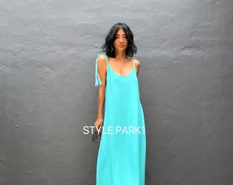 May include: A long, turquoise maxi dress with thin shoulder straps and a ruffled hem. The dress has a flowing silhouette and is made of a lightweight fabric. The text "STYLE PARK1" is visible on the dress. The model is wearing the dress in front of a gray wall.