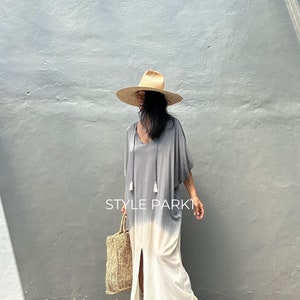 May include: A woman wearing a gray and white tie-dye maxi dress with a side slit and tassels. She is also wearing a wide-brimmed straw hat and sandals. The dress has a V-neckline and is made of a lightweight fabric. The woman is holding a woven tote bag. The text "STYLE PARK1" is visible in the image.
