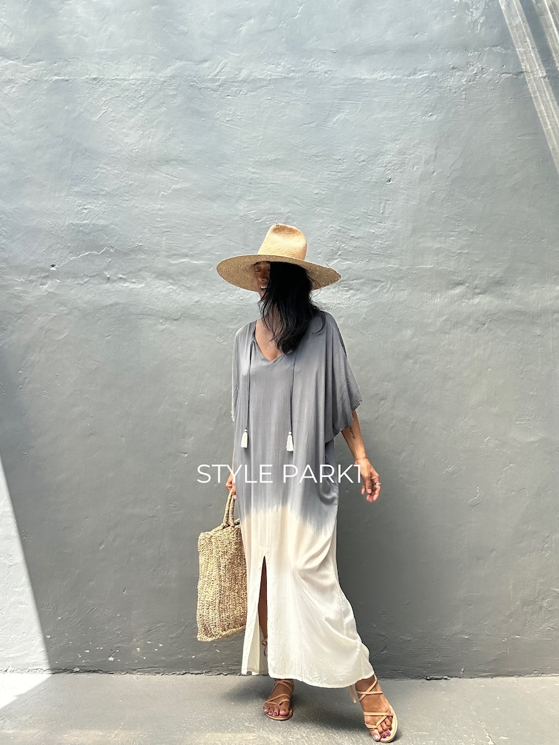 May include: A woman wearing a gray and white tie-dye maxi dress with a side slit and tassels. She is also wearing a wide-brimmed straw hat and sandals. The dress has a V-neckline and is made of a lightweight fabric. The woman is holding a woven tote bag. The text "STYLE PARK1" is visible in the image.