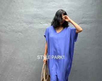 May include: A long, flowing, periwinkle blue dress with a V-neck and short sleeves. The dress is paired with a woven beige tote bag. The text "STYLE PARK1" is visible on the dress. The background is a solid gray wall.