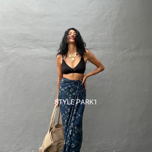 Puede incluir: Una mujer con un sujetador negro y una falda de envoltura con estampado azul y blanco. Lleva un bolso de mano tejido y un collar de oro con un colgante.