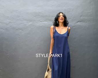 May include: A woman wearing a navy blue maxi dress with thin straps that tie at the shoulders. The dress has a ruffled hem and the woman is holding a woven tote bag.