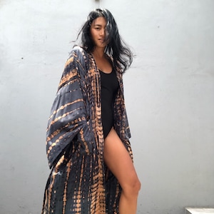May include: A long, flowing kimono-style robe in a tie-dye pattern of black and tan. The robe has wide sleeves and is open in the front. The model is wearing a black bodysuit underneath.