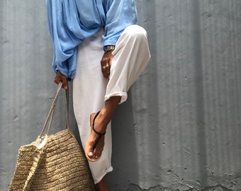 May include: A light blue blouse with long sleeves and a button-down front. The outfit includes white pants, black sandals, and a large woven tote bag. The bag is made of a light brown material.