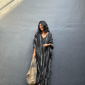 May include: A black and white patterned caftan dress with a V-neck. The dress features vertical white stripes on a black background. A beige woven tote bag is held. Brown sandals are worn.