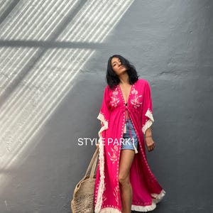 May include: A pink maxi dress with white embroidery and fringe detailing. The dress has a V-neckline and a tie at the waist. The model is wearing the dress with denim shorts and a straw tote bag. The text "STYLE PARK1" is visible in the image.