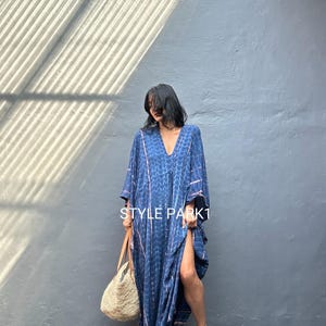May include: A long, flowing blue dress with a V-neck and a high slit. The dress has a patterned design and is paired with a straw tote bag and sandals. The text "STYLE PARK1" is visible on the dress.