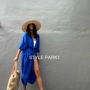 Puede incluir: Una mujer lleva un kimono azul real con una prenda interior blanca, un sombrero de paja de ala ancha y tacones altos dorados. Lleva un bolso de mano tejido y está de pie frente a una pared gris. El texto "STYLE PARK1" es visible en la pared.