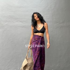 Puede incluir: Una mujer lleva un top de sujetador negro y una falda de sarong con estampado morado y naranja. Lleva un bolso de mano de crochet beige. El texto "STYLE PARK1" es visible en la imagen.