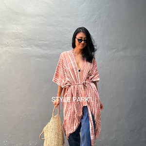 May include: A woman wearing a pink and white tie-dye kimono with a brown belt, blue jeans, and sunglasses. She is carrying a woven straw tote bag. The text "STYLE PARK1" is visible in the image.