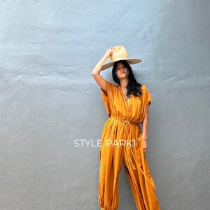 May include: A woman wearing a mustard yellow jumpsuit with a wide-brimmed straw hat. The jumpsuit has a cinched waist with a rope belt and a relaxed fit. The woman is standing in front of a gray wall with the text "STYLE PARK" printed on it.