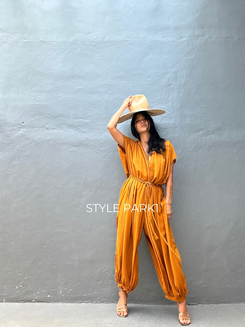 May include: A woman wearing a mustard yellow jumpsuit with a wide-brimmed straw hat. The jumpsuit has a cinched waist with a rope belt and a relaxed fit. The woman is standing in front of a gray wall with the text "STYLE PARK" printed on it.