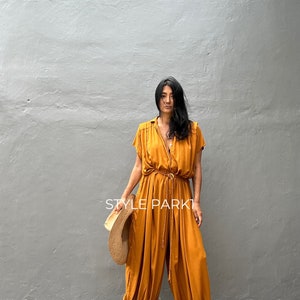 May include: A woman wearing a mustard yellow jumpsuit with a cinched waist and a wide leg. She is holding a straw hat and is standing in front of a gray wall. The text "STYLE PARK1" is visible in the image.