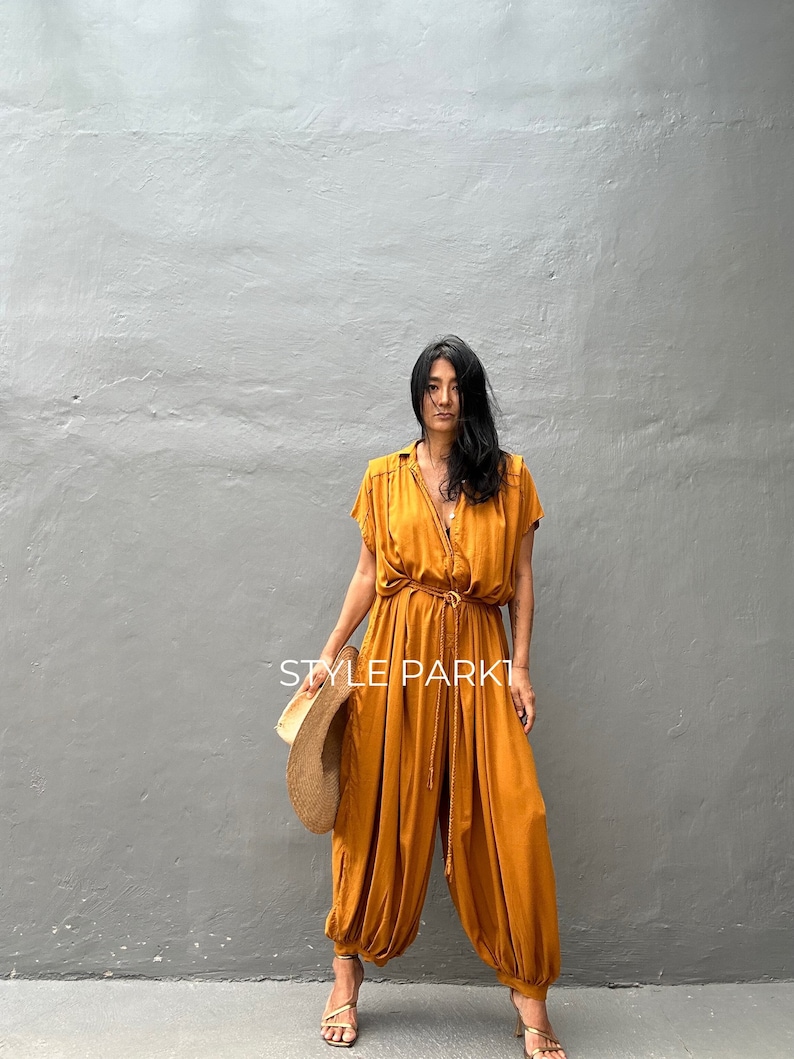 May include: A woman wearing a mustard yellow jumpsuit with a cinched waist and a wide leg. She is holding a straw hat and is standing in front of a gray wall. The text "STYLE PARK1" is visible in the image.