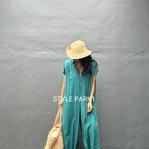 Peut inclure: Une combinaison bleu sarcelle avec une fermeture boutonn&eacute;e sur le devant, des manches courtes et des chevilles resserr&eacute;es. Le v&ecirc;tement porte le texte STYLE PARK1. Elle est associ&eacute;e &agrave; un chapeau de paille, un grand sac fourre-tout en osier et des sandales.