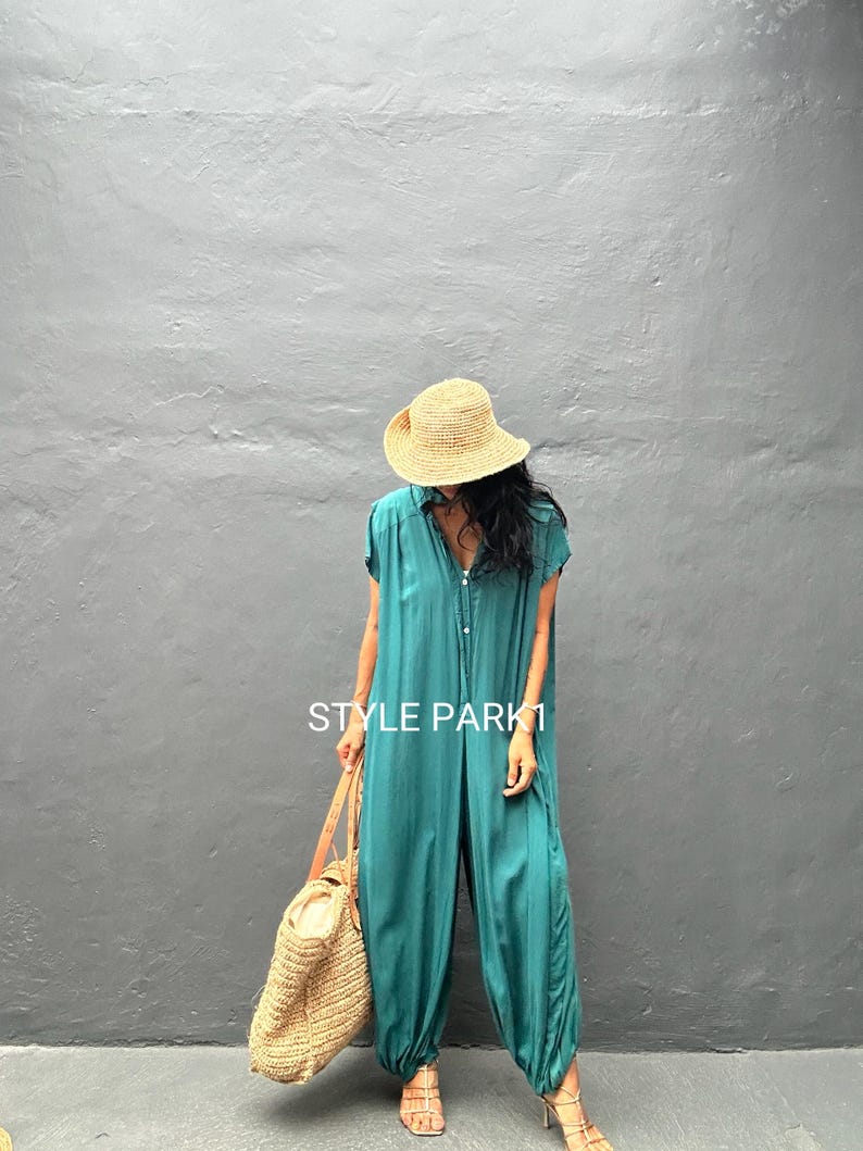 Peut inclure: Une combinaison bleu sarcelle avec une fermeture boutonn&eacute;e sur le devant, des manches courtes et des chevilles resserr&eacute;es. Le v&ecirc;tement porte le texte STYLE PARK1. Elle est associ&eacute;e &agrave; un chapeau de paille, un grand sac fourre-tout en osier et des sandales.