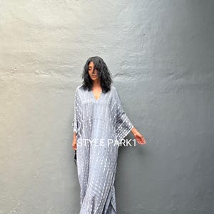 May include: A gray and white tie-dye kaftan with a V-neck and long sleeves. The kaftan is loose-fitting and has a flowing silhouette. The model is wearing the kaftan with sandals. The text "STYLE PARK1" is visible on the bottom of the image.